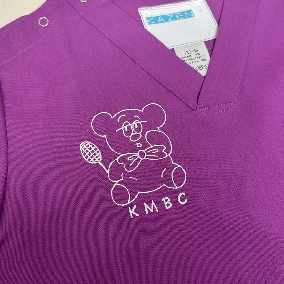 パープル地のスクラブに施された、北里大学医学部バドミントン部（KMBC）の白いキャラクター刺繍。ラケットを持ち蝶ネクタイをしたクマの線画デザイン。
