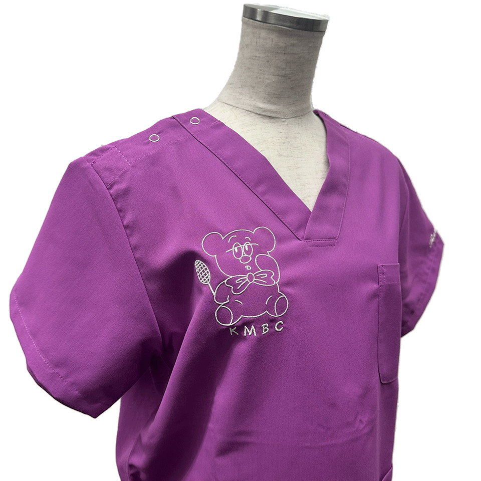 パープル地のスクラブに施された、北里大学医学部バドミントン部（KMBC）の白いキャラクター刺繍。ラケットを持ち蝶ネクタイをしたクマの線画デザイン。