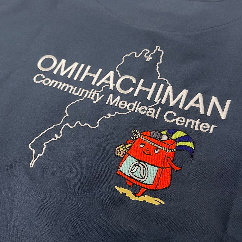 スクラブ背面の刺繍正面アップ。白の線地図で描かれた琵琶湖を背景に、「OMIHACHIMAN Community Medical Center」の文字と赤コン君が刺繍されている。
