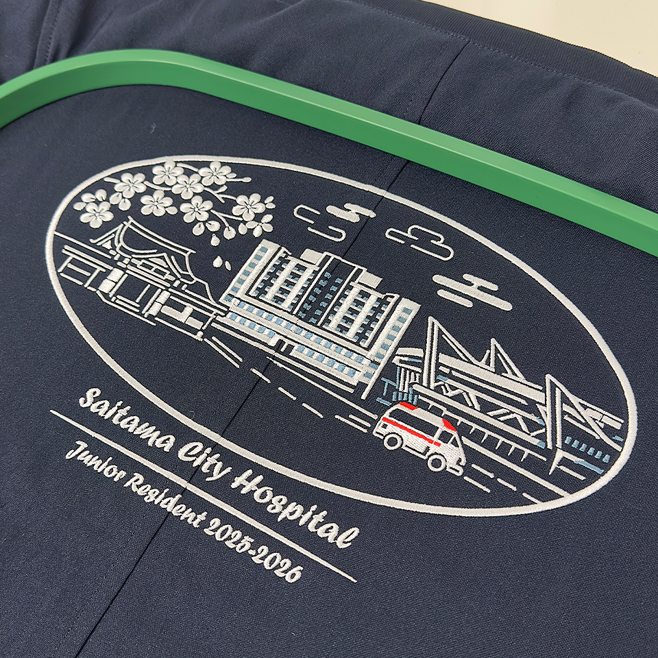 紺色のブルゾン背面に施された、さいたま市立病院「初期研修医 2025-2026」記念刺繍の全体像。さいたま市の街並みが楕円の中にデザインされている。