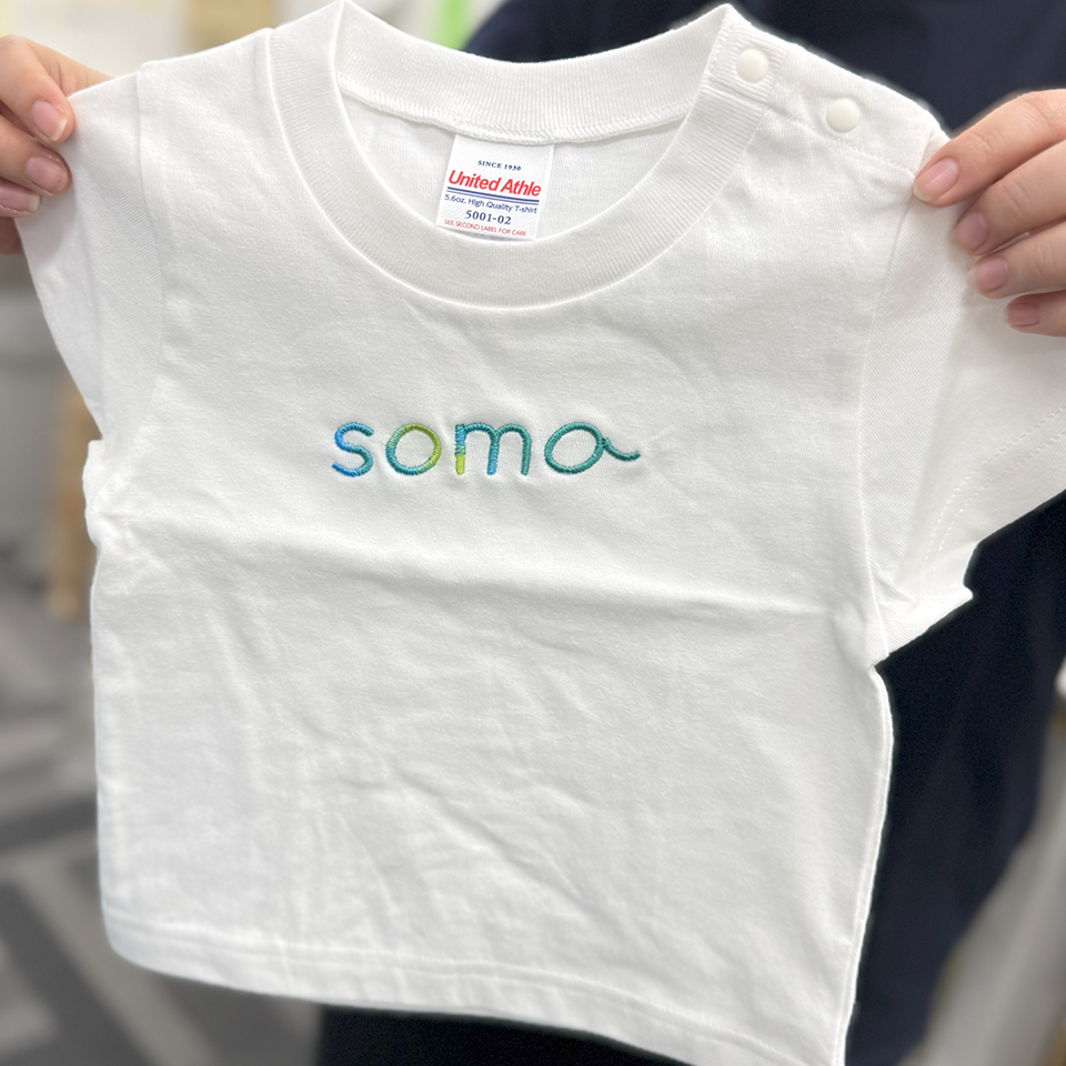 「soma」という文字のグラデーション刺繍の拡大写真。サックスブルーからイエローグリーン、エメラルドグリーンへと滑らかに色が移り変わる糸の質感とステッチの詳細。